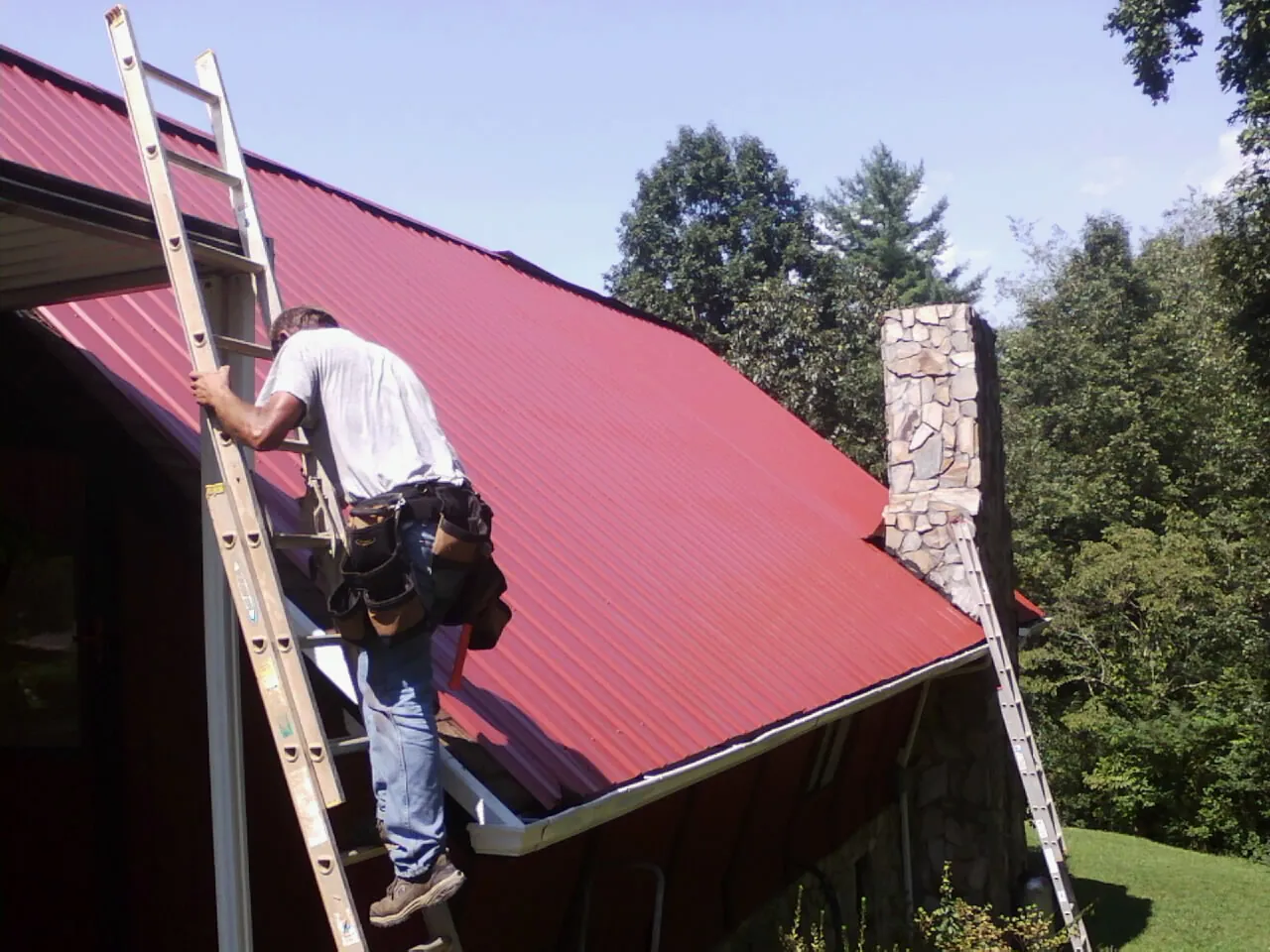 Roofer on a ladder beside a red metal roof and stone chimney — Soffit & Fascia Repair by Roofing Brothers in Calhoun